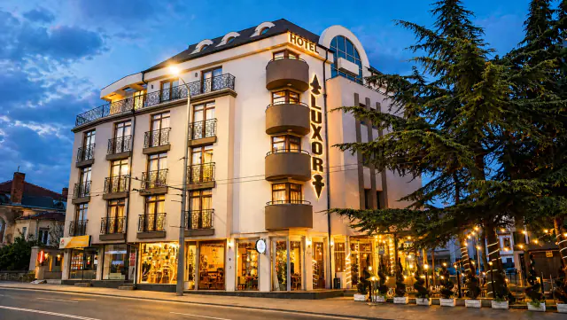 Fasáda hotelu Luxor za soumraku s osvětleným nápisem LUXOR a letní zahrádkou se světelnými girlandami.