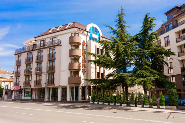 Hotel Luxor v ulici Bulair za slunečného dne, s cedry před budovou.